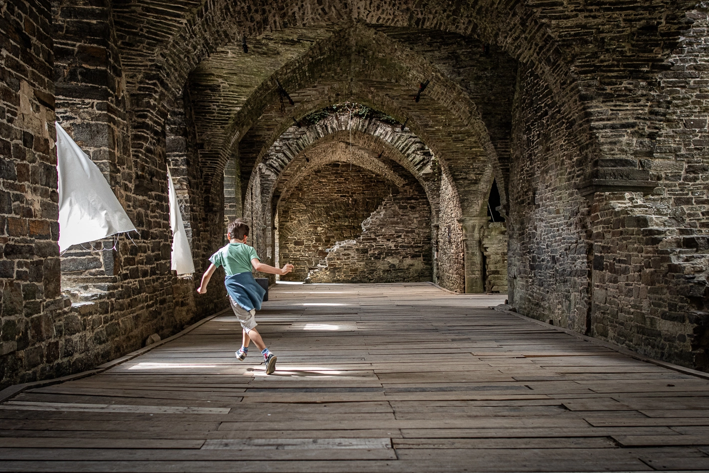 Garçon qui joue dans une pièce. Il semble courir. La pièce est lumineuse. Il y a des faisceaux de lumières qui passent dans les arches.