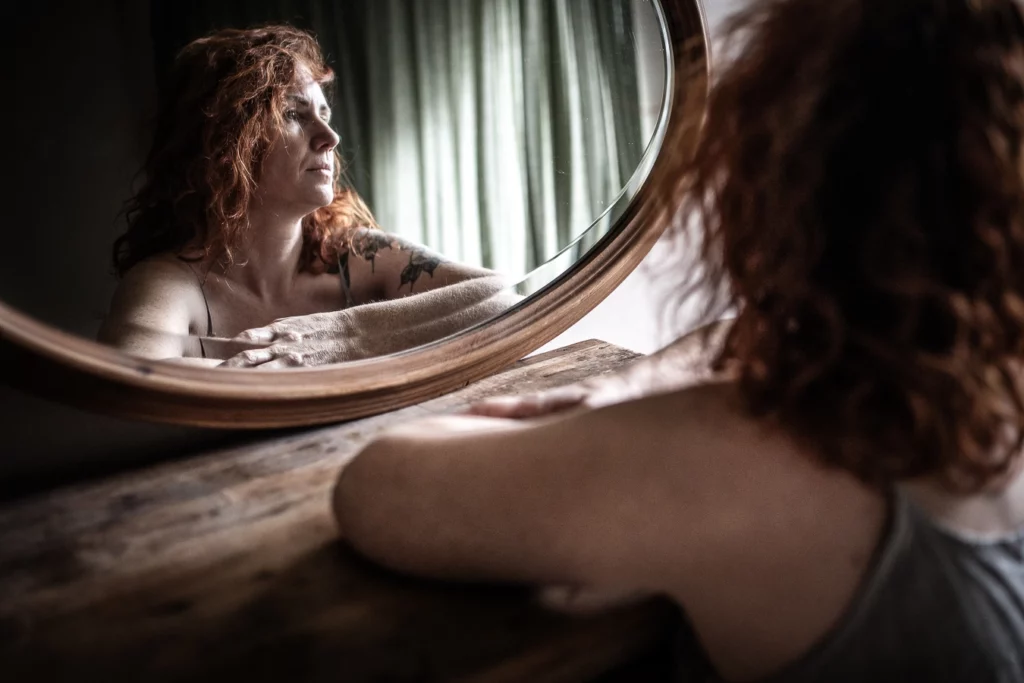 Femme qui regarde son reflet dans un miroir en étant appuyée sur un bureau. Il y a des faisceaux de lumière qui passent à travers les rideaux. La pièce semble sombre et lumineuse à la fois. Elle a aussi un tatouage. Boudoir