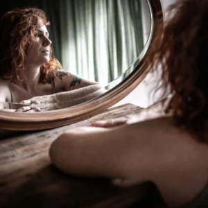 Femme qui regarde son reflet dans un miroir en étant appuyée sur un bureau. Il y a des faisceaux de lumière qui passent à travers les rideaux. La pièce semble sombre et lumineuse à la fois. Elle a aussi un tatouage. Boudoir