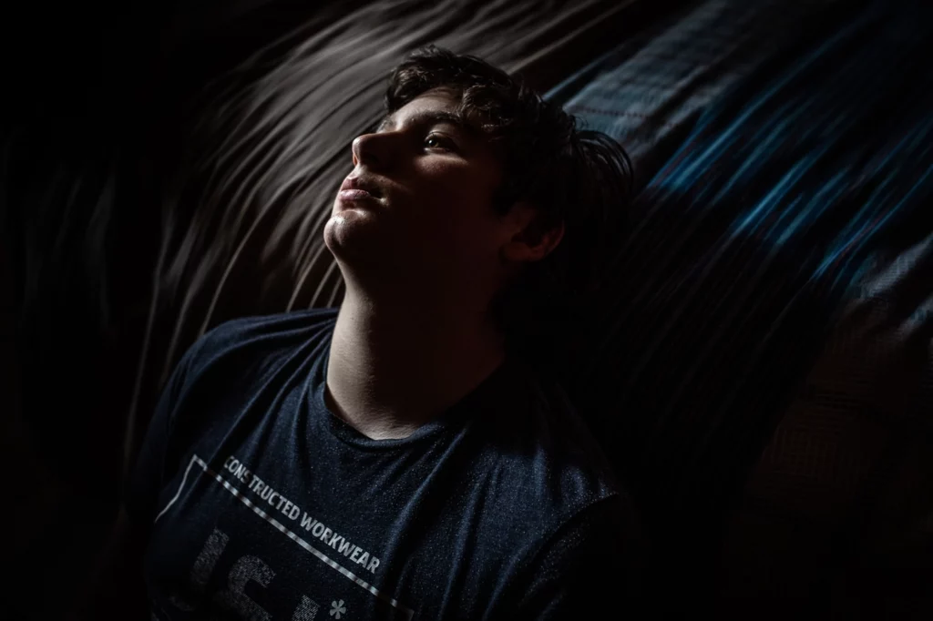 Un rayon de lumière éclaire le visage d'un garçon assis sur le sol qui repose sa tête contre son lit. Il porte un t-shirt bleu.
