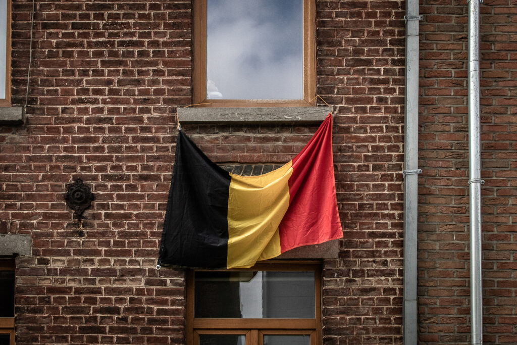 Un drapeau aux couleurs nationales belges pend sur une fenêtre. La façade est faite de briques rouges. Les chassis de la maison sont bruns et le ciel reflète dans la fenêtre.