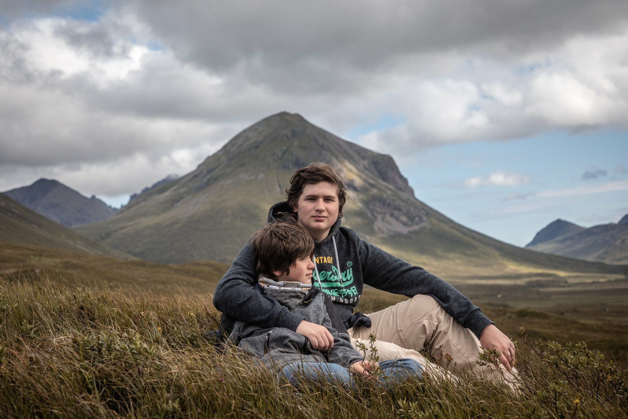 Enfants au milieu d'un champ devant des montages et un ciel nuageux. Ils sont assis.
