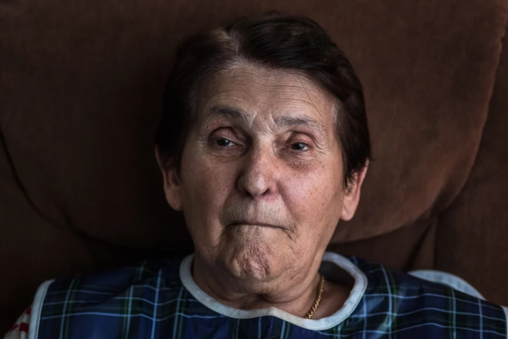 Femme qui semble sourire. Elle porte un haut bleu et est contre un mur noir.